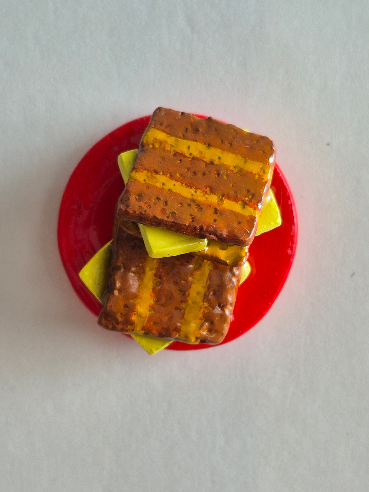 A miniature kaya toast magnet. Brown striped bread, with yellow butter slices between. 2 such toast stacked on each other, on a small red plate on a light gray background.