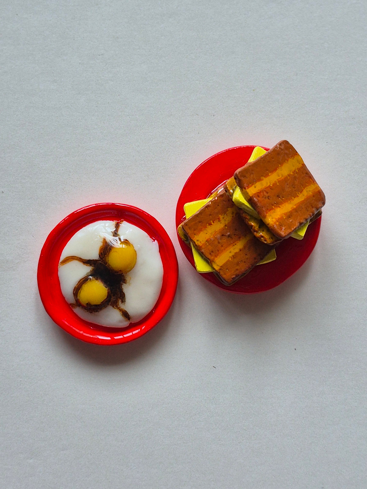 Two miniature magnets against a light gray background. 2 kaya toast bread on a circular red plate. 2 soft-boiled eggs with black sauce on a red circular plate.